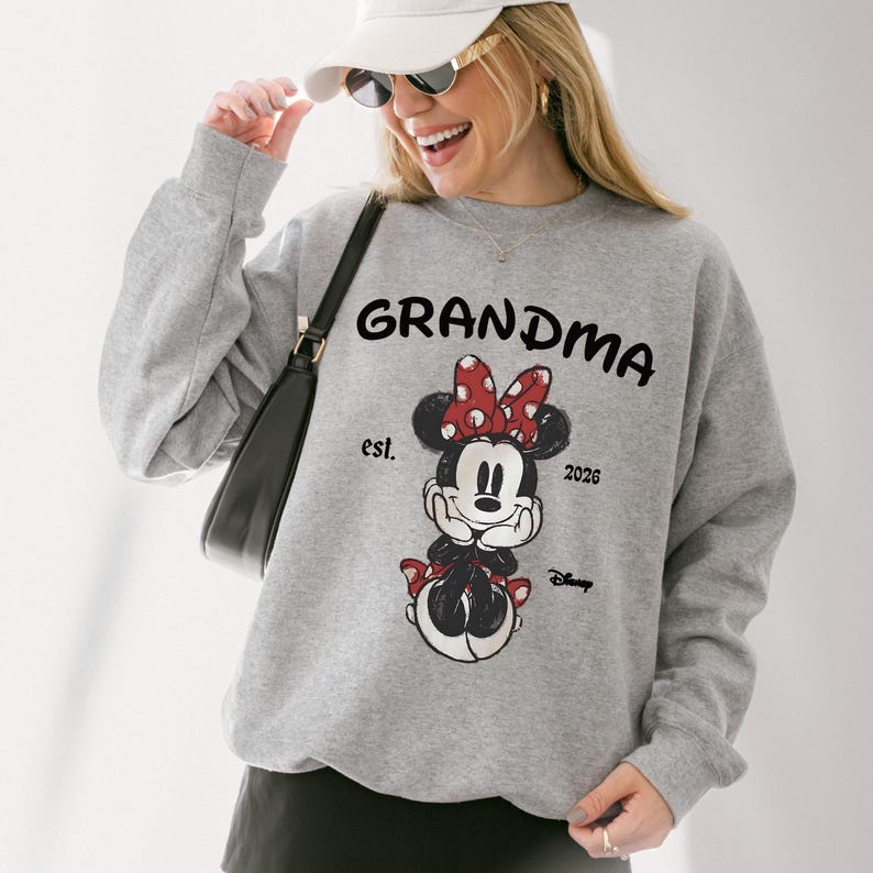 Op de afbeelding: Een lichtgrijs sweatshirt met het woord "GRANDMA" in zwarte blokletters. Daaronder een Minnie Mouse-afbeelding met een rode strik en de tekst "est. 2026". Het Disney-logo staat rechtsonder.