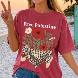 May include: A maroon t-shirt with the text "Free Palestine" in white. The shirt features a floral design with red poppies, white daisies, and a black and white scarf pattern. The shirt has short sleeves.