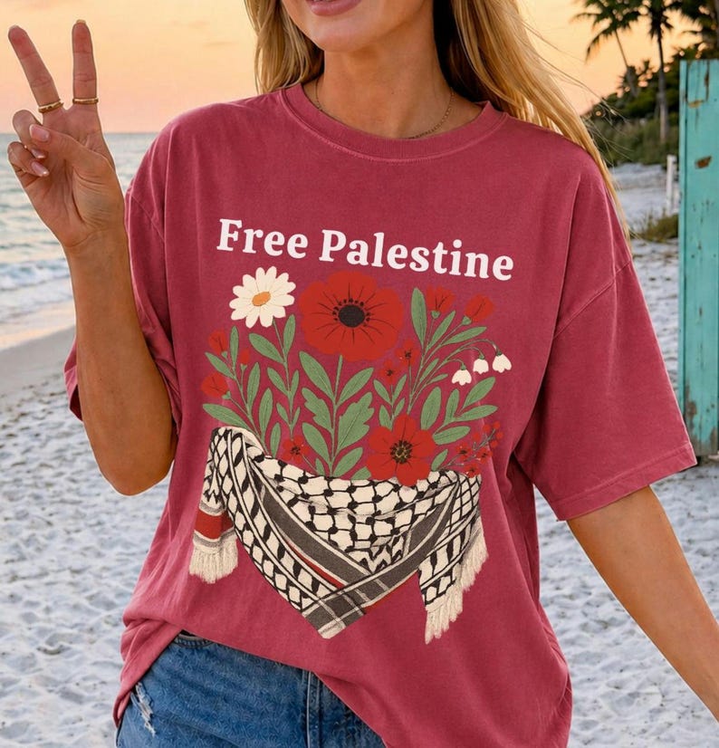 May include: A maroon t-shirt with the text "Free Palestine" in white. The shirt features a floral design with red poppies, white daisies, and a black and white scarf pattern. The shirt has short sleeves.