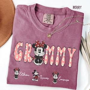 May include: Berry-colored t-shirt with the word "Grammy" in a floral design, featuring a Minnie Mouse graphic. The shirt includes the names "Ethan", "Lucas", and "Emma" below the graphic. A gold necklace is draped across the shirt.