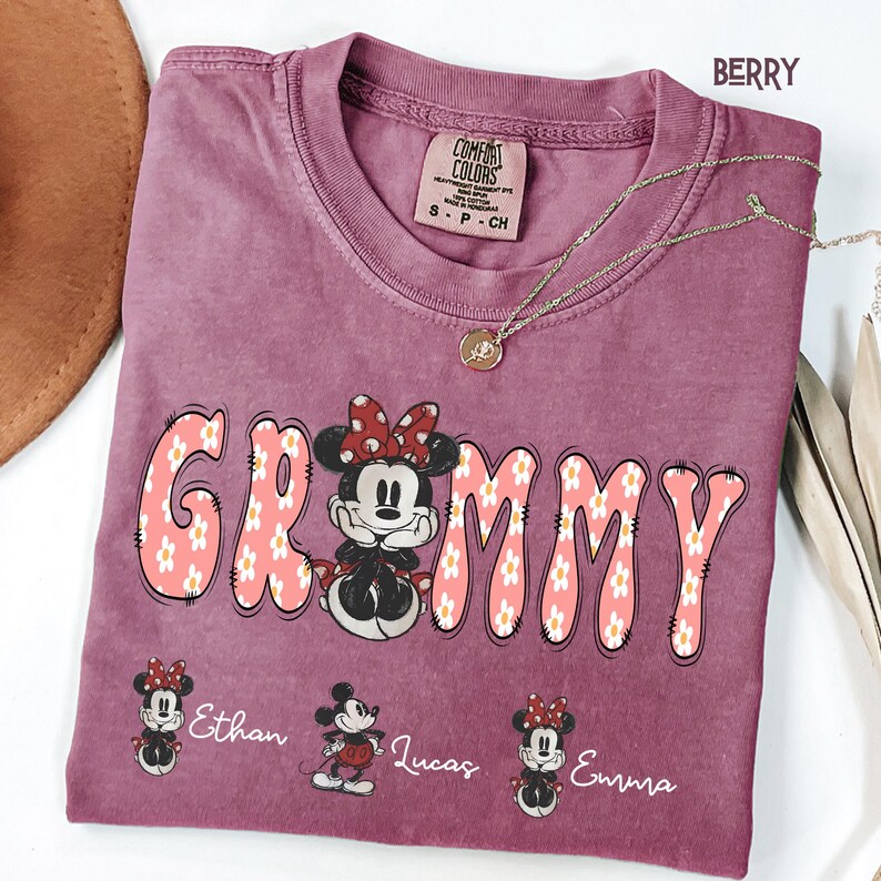 May include: Berry-colored t-shirt with the word "Grammy" in a floral design, featuring a Minnie Mouse graphic. The shirt includes the names "Ethan", "Lucas", and "Emma" below the graphic. A gold necklace is draped across the shirt.