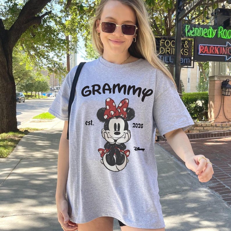 K&ouml;nnte beinhalten: Hellgraues T-Shirt mit dem Wort "GRAMMY" in schwarzer Schrift &uuml;ber einer Minnie Mouse-Grafik. Die Grafik enth&auml;lt eine rote Schleife mit wei&szlig;en Punkten, und der Text "est. 2026" und "Disney" sind ebenfalls vorhanden. Das T-Shirt ist l&auml;ssig.