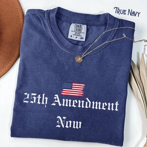 May include: A navy blue t-shirt with the text "25th Amendment Now" in white gothic font, and a small American flag graphic. The shirt has a crew neck and short sleeves. A gold necklace with a pendant is draped across the shirt.