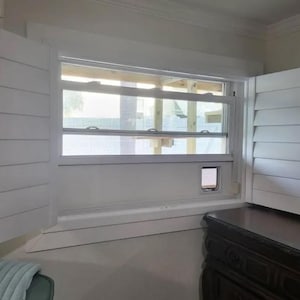 May include: A white window frame with horizontal louvered shutters on both sides. The window features a built-in rectangular pet door integrated into the lower right section of the frame. The glass pane shows a view of an outdoor structure.