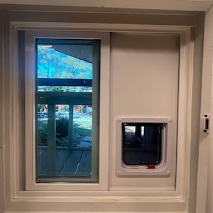 May include: A white-framed window with a sliding glass panel and a pet door installed in the adjacent wall. The pet door has a white frame and a clear flap. The window offers a view of a backyard.