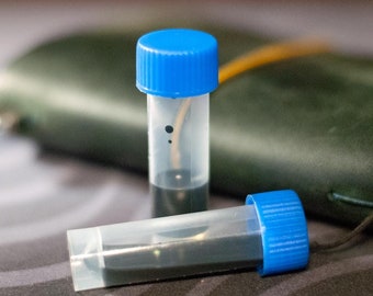 Puede incluir: Dos pequeños frascos de plástico transparente con tapas de rosca azules llenos de tinta negra. Los frascos están sobre una superficie gris oscuro con un cuaderno de cuero verde en el fondo.