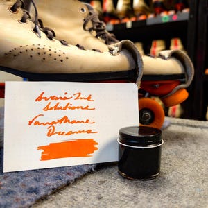 May include: A small jar of orange ink with the words "Inks Ink Solutions Vanathane Dreams" written on a white card. The ink is a vibrant orange colour and the jar is black.