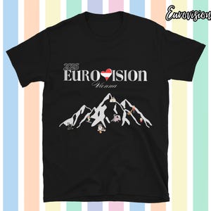 Op de afbeelding: Zwart T-shirt met de tekst "2026 EUROVISION Vienna" en een hartvormige afbeelding in de kleuren van de Oostenrijkse vlag. Daaronder een bergketen met kleine figuren. Het T-shirt heeft korte mouwen.