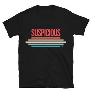 Op de afbeelding: Zwart t-shirt met het woord "SUSPICIOUS" in rood, boven horizontale strepen in oranje, beige, turquoise en blauw. Het shirt heeft een ronde hals en korte mouwen.
