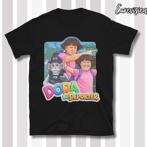 May include: Black t-shirt featuring a humorous graphic of a cartoon character with the text "DORA the DEPORTER". The graphic includes a monkey wearing an "ICE" vest and two figures in pink shirts and backpacks.