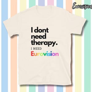 Op de afbeelding: Crèmekleurig T-shirt met de tekst "I don't need therapy. I need Eurovision" in zwart en regenboogkleuren. Het shirt heeft een ronde hals en korte mouwen. De achtergrond is gestreept met pastelkleuren.