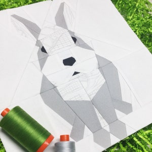 Op de afbeelding: Een wit stoffen paneel met een grijs geometrisch konijnontwerp. Het konijn is gemaakt van veel kleine driehoeken. Er liggen twee spoelen garen op de stof, een groene en een grijze.