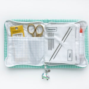 Puede incluir: Una bolsa con cremallera de cuadros verdes y blancos con un pequeño amuleto de coche. La bolsa está abierta y contiene un pequeño par de tijeras doradas, un paquete de agujas etiquetadas como "hidamari" y una tela blanca con la palabra "TAPESTRY" y una fila de agujas.