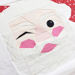May include: A white fabric patchwork quilt block with a Santa Claus face design. The face is made of white, pink, and black fabric pieces. The Santa has a red hat with white polka dots.