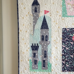 Puede incluir: Una colcha blanca con un diseño de castillo azul y gris. El castillo tiene una bandera roja y está rodeado de estrellas doradas. La colcha está hecha de algodón y es perfecta para la habitación de un niño.