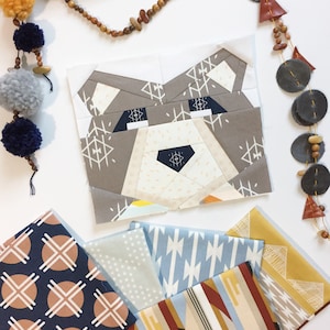 May include: A collection of fabric squares with geometric patterns in shades of blue, brown, white, and yellow. The fabric squares are arranged in a stack, with the top square featuring a bear face design.