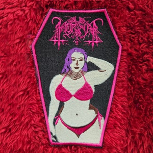 May include: A black and pink embroidered patch in the shape of a coffin. The patch features a woman in a pink bikini with the word "HORNS" in pink lettering above her.