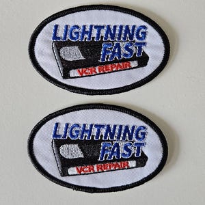 May include: Two oval embroidered patches with a white background and black border. Each patch features a black VHS tape graphic with the blue text LIGHTNING FAST and the red text VCR REPAIR embroidered across the center of the design.