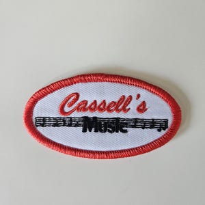 May include: An oval-shaped embroidered patch with a white background and a red border. The text 'Cassell's' is stitched in red script above the word 'Music' in black block letters, which is superimposed over a black musical staff line.