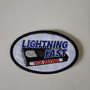 May include: An oval-shaped embroidered patch with a white background and a black border. It features a black VHS tape graphic with the blue text LIGHTNING FAST and the red text VCR REPAIR embroidered across the center of the design.
