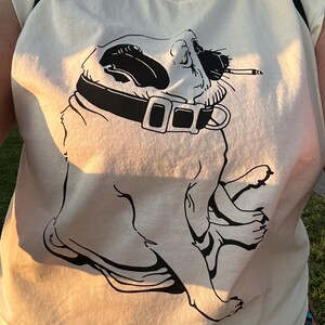 May include: A cream-colored t-shirt featuring a black graphic print of a pug wearing a collar and holding a cigarette in its mouth. The dog is depicted in profile, sitting down with its head tilted back, rendered in a bold, minimalist line art style.
