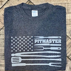 May include: Dark gray t-shirt with a white graphic of the American flag, with the stars replaced by stars and the stripes replaced by grilling tools. The word "PITMASTER" is printed in white.