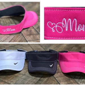 May include: Three visors, one white, one black, and one pink. The pink visor has the word "Mom" embroidered on it with a heart symbol. All three visors have a white tick logo on the side.