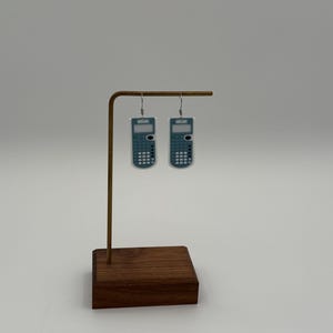May include: A pair of calculator-shaped earrings. The earrings are teal and white, with a rectangular shape and a small screen at the top. The earrings are hanging from a gold-colored metal stand with a wooden base.