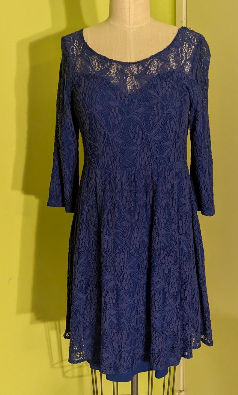 Op de afbeelding: Een marineblauwe kanten jurk met lange mouwen en een ronde halslijn. De jurk wordt gepresenteerd op een mannequin en heeft een getailleerd lijfje dat uitloopt in een volle rok. De ingewikkelde kanten details bedekken de hele jurk.