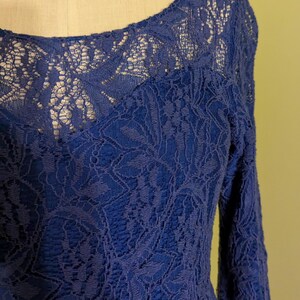 Op de afbeelding: Een kobaltblauwe kanten jurk met lange mouwen. De jurk heeft een ronde hals en ingewikkelde bloemenkanten details over de hele bodice en mouwen. De jurk wordt op een mannequin getoond.