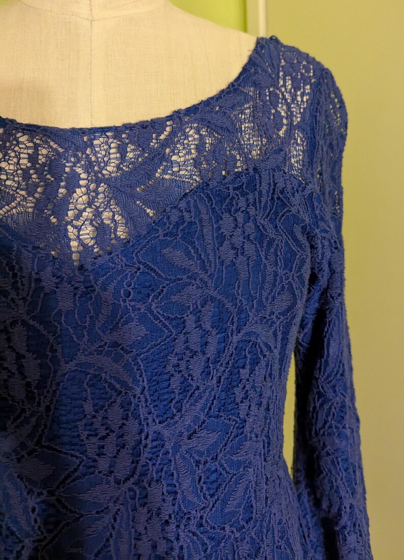 Op de afbeelding: Een kobaltblauwe kanten jurk met lange mouwen. De jurk heeft een ronde hals en ingewikkelde bloemenkanten details over de hele bodice en mouwen. De jurk wordt op een mannequin getoond.