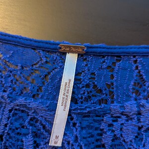 Op de afbeelding: Close-up van een koningsblauwe kanten top. De top heeft een delicaat bloemen kanten patroon. Een label met de merknaam "Free People" is zichtbaar, samen met een label met de tekst "Made in China" en "Fabriqu&eacute; en Chine". De maat is medium.
