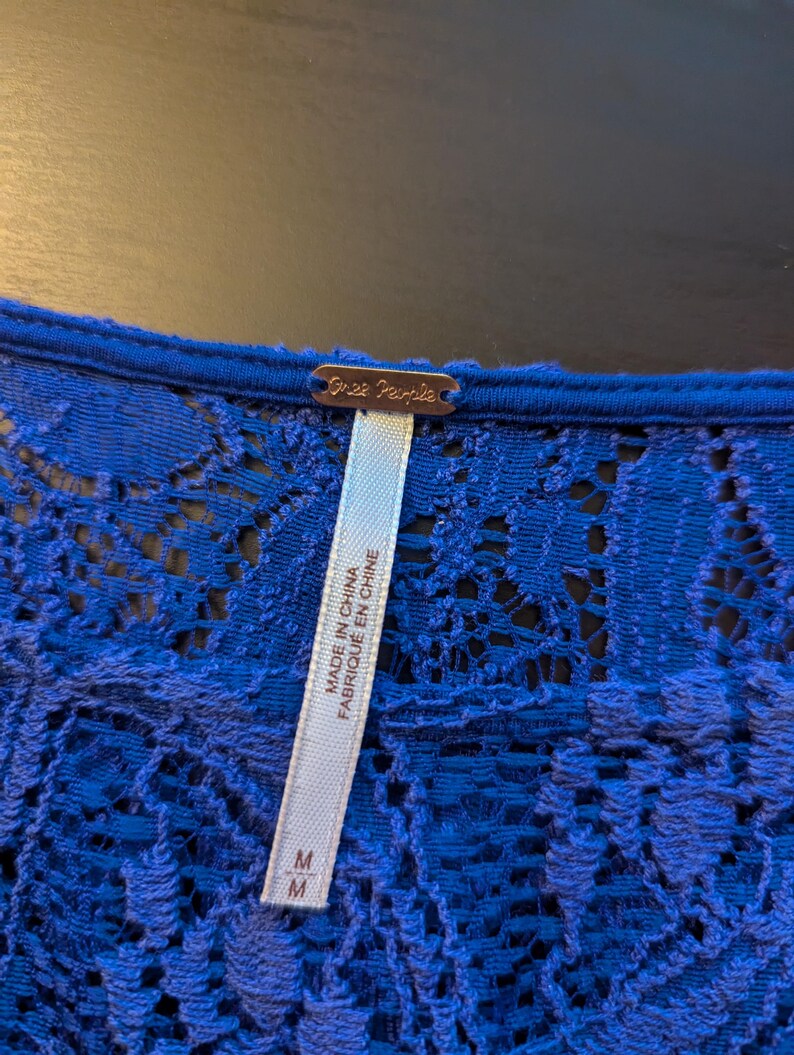 Op de afbeelding: Close-up van een koningsblauwe kanten top. De top heeft een delicaat bloemen kanten patroon. Een label met de merknaam "Free People" is zichtbaar, samen met een label met de tekst "Made in China" en "Fabriqu&eacute; en Chine". De maat is medium.