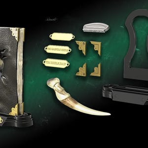 Puede incluir: Un libro encuadernado en cuero negro con lomo dorado y una garra que sobresale de la portada. El libro está sobre un soporte negro con una placa dorada que dice "The Journal of the First Riddle". El libro forma parte de un conjunto de accesorios de la película "The Ritual".