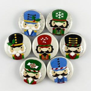 May include: Seven circular Christmas-themed badges with cartoon nutcracker soldier illustrations. Each badge has a different colour scheme, including blue, green, red, and black, with details like holly, snowflakes, and candy canes.