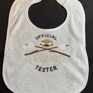 May include: White baby bib with brown embroidery that says "OFFICIAL SMORE TESTER" with a graphic of a s'more and crossed sticks.