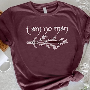 May include: Burgundy t-shirt with the text "I am no man" in a white, elegant font, accompanied by a sword and floral design. The shirt is folded at the bottom. A white mug and woven mat are in the background.
