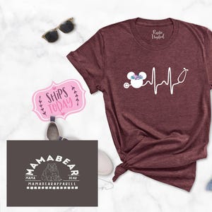 May include: A maroon t-shirt with a white graphic of a Mickey Mouse head with a floral crown and a stethoscope, with a heartbeat line and the text "Ships Today".