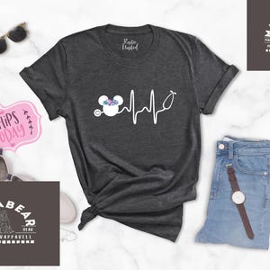 May include: A dark grey t-shirt with a white graphic of a Mickey Mouse head wearing a floral crown and a stethoscope with a heartbeat line. The text "Mama Bear" is on a dark grey rectangle with white text.