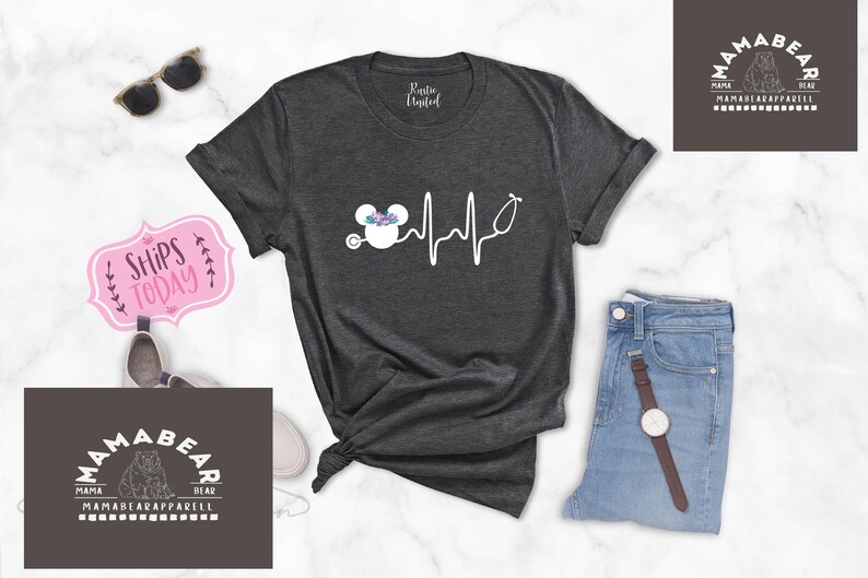 May include: A dark grey t-shirt with a white graphic of a Mickey Mouse head wearing a floral crown and a stethoscope with a heartbeat line. The text "Mama Bear" is on a dark grey rectangle with white text.