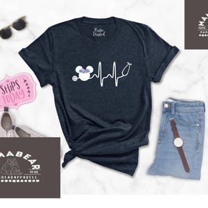 May include: A navy blue t-shirt featuring a white outline of a Mickey Mouse head with flowers on top and a stethoscope. The Mickey Mouse head is connected to a white heartbeat line. The text "Mama Bear" is on a brown rectangle in the top right corner of the image. The text "Mama Bear Apparel" is on a brown rectangle in the bottom left corner of the image.