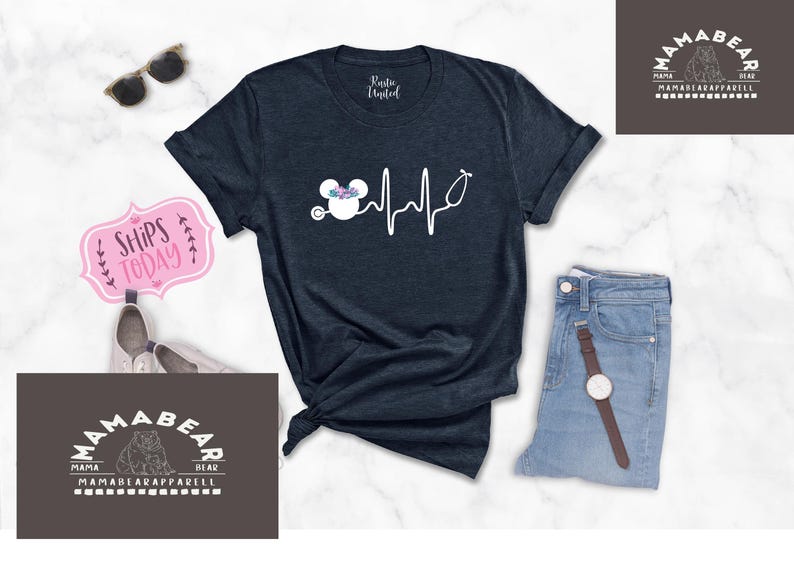 May include: A navy blue t-shirt featuring a white outline of a Mickey Mouse head with flowers on top and a stethoscope. The Mickey Mouse head is connected to a white heartbeat line. The text "Mama Bear" is on a brown rectangle in the top right corner of the image. The text "Mama Bear Apparel" is on a brown rectangle in the bottom left corner of the image.