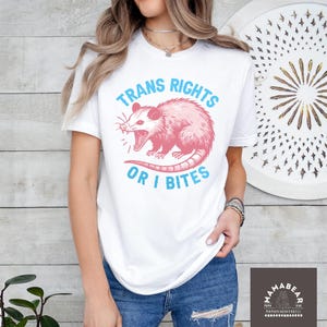 May include: A white short-sleeve t-shirt featuring a graphic of a pink opossum with its mouth open. The text "TRANS RIGHTS OR I BITES" is printed in light blue capital letters above and below the opossum illustration on the front of the shirt.