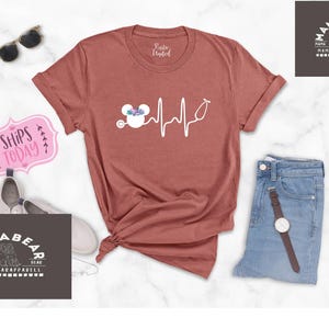 May include: A rust-coloured t-shirt with a white graphic of a Mickey Mouse head with flowers and a stethoscope, and a heartbeat line. The text "Ships Today" is on a pink sign in the background.
