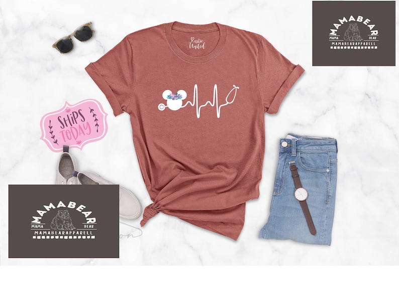 May include: A rust-coloured t-shirt with a white graphic of a Mickey Mouse head with flowers and a stethoscope, and a heartbeat line. The text "Ships Today" is on a pink sign in the background.