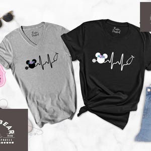 May include: Two grey and black t-shirts with a Mickey Mouse design and a heartbeat graphic. The graphic includes a stethoscope and a flower crown on Mickey Mouse's head. The text on the shirts reads "Ships Today".
