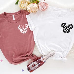 May include: Two t-shirts with a checkered Mickey Mouse design. One shirt is white and the other is a dusty rose color.