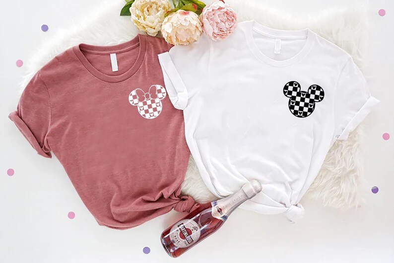 May include: Two t-shirts with a checkered Mickey Mouse design. One shirt is white and the other is a dusty rose color.