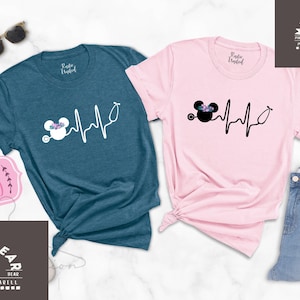 May include: Two t-shirts featuring a black and white graphic of a cartoon mouse with a stethoscope and a heartbeat line. The t-shirts are teal and pink.