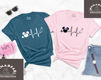 May include: Two t-shirts with a black and white graphic of a cartoon mouse with a stethoscope and a heartbeat line. The t-shirts are teal and pink.